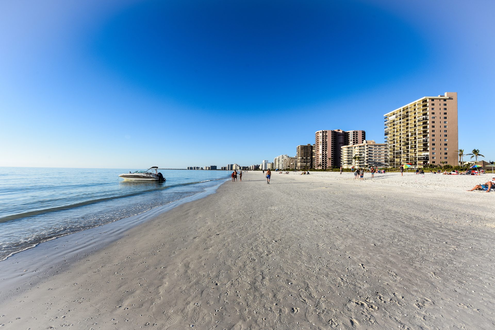 A boat pulled up on a wide sandy Marco Island beach under a deep blue sky, with beachfront condominium towers in the distance