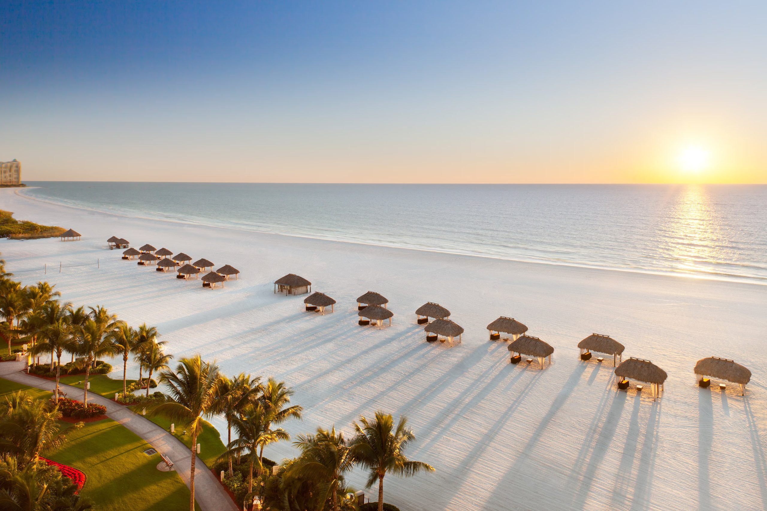 Sunrise over the Gulf and a wide white sand beach on Marco Island, with rows of beach umbrellas and palms along the shore