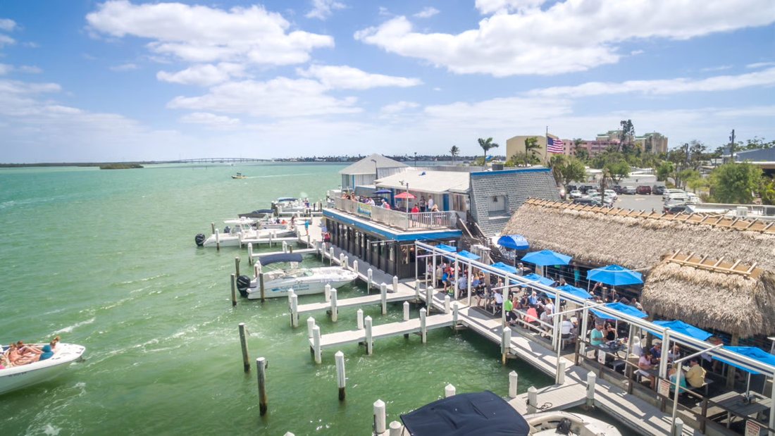 Aerial view of a waterfront restaurant with boat docks, blue umbrellas and a thatched roof on the Marco River